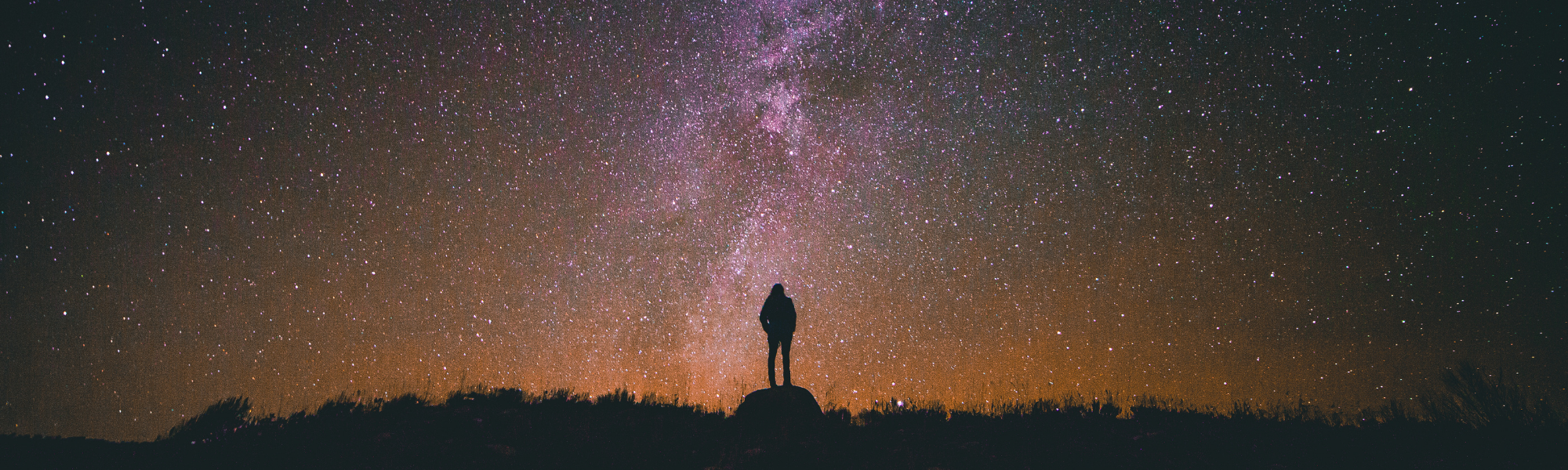 Image shows a person standing on a ridge looking out into the night sky which is filled with stars and the Milky Way.  Image courtesy of Canva