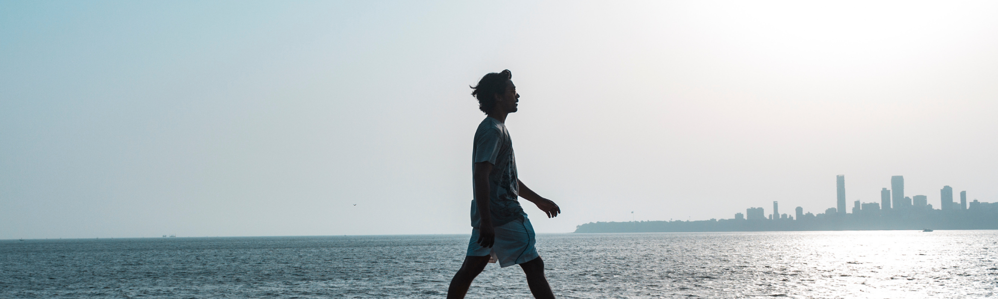 Image shows a young man walking along an ocean harbour with the city skyline in the background.  Image courtesy of Canva