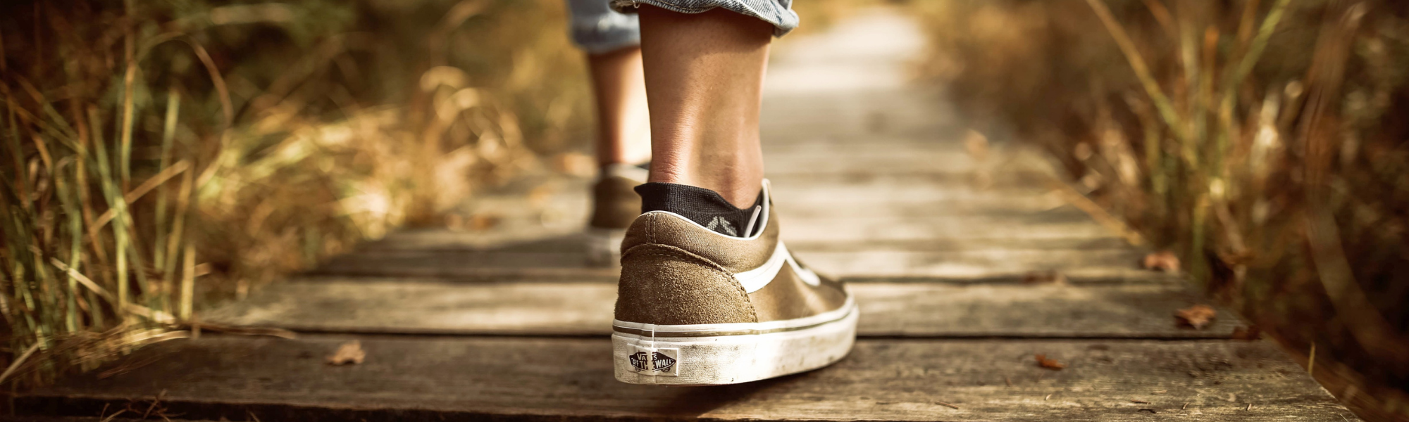 Image shows a lady wearing tan sneakers walking on a boardwalk surrounded by grasses.  Image courtesy of Canva