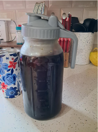 A large glass container with a gray lid and handle, filled with dark liquid, likely cold brew coffee, sitting on a speckled kitchen countertop. In the background are utensils, a floral tumbler, a white container with red-handled knives, and a yellow item.